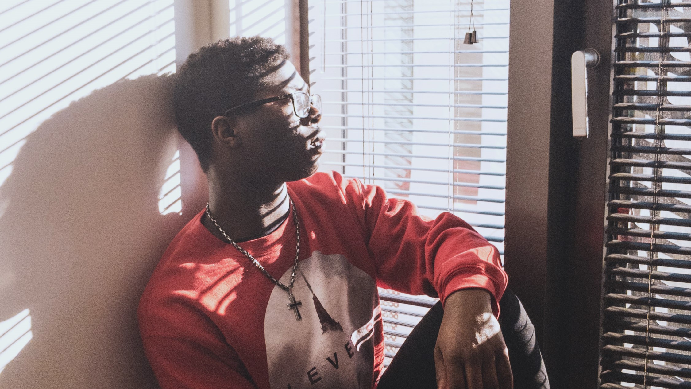 picture of Emmanuel sitting on a windowsill in a 
                        red sweatshirt looking out the window